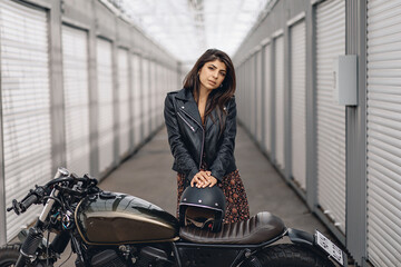 Fototapeta na wymiar Young adorable beautiful girl in a leather jacket and dress posing next to a motorcycle and holding a black helmet in her hands and looking straight at the camera. Extreme hobby concept