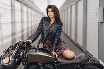Fototapeta na wymiar Portrait of a young and beautiful lady in a black leather jacket and dress posing next to a black retro motorcycle and looking playfully straight into the camera. Sensuality concept
