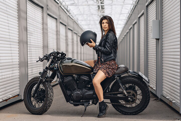 Naklejka na ściany i meble Girl on a motorcycle. Lovely brave female driver in a leather jacket and dress sitting on her retro motorcycle and holding a helmet in her hands is getting ready to go. Speed ​​concept