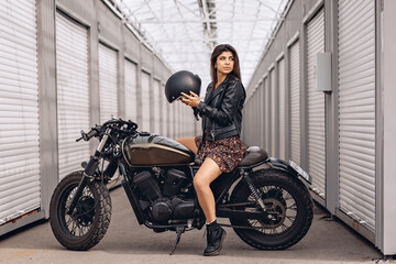 Fototapeta na wymiar Girl on a motorcycle. Beautiful female driver in a leather jacket and dress sitting on her retro motorcycle and holding a helmet in her hands is getting ready to go. Moto ​​concept