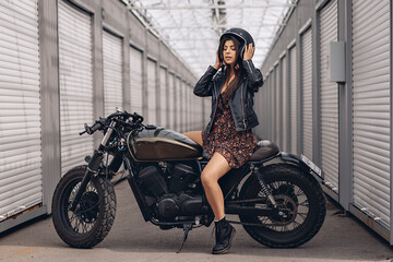 Naklejka na ściany i meble Beautiful young biker woman in a leather jacket and dress sitting on a motorcycle and with a helmet on her head with her eyes closed on a white background. Extreme concept