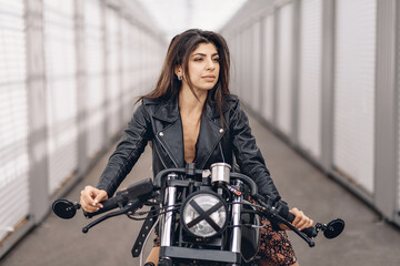 Fototapeta na wymiar Portrait of a sexy attractive dark-haired driver girl in a black leather jacket sitting on a black retro motorcycle and looking straight into the distance against a white background. Extreme concept