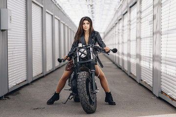 Lovely young and brutal biker movel in a leather jacket posing on a black retro motorcycle holding the steering wheel with her hands and looking into the distance. Extreme concept