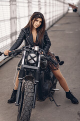 Fototapeta na wymiar Portrait of a sexual dark-haired lady in a black leather jacket sitting and posing on a black retro motorcycle against a white background of walls and streets. Brutality concept