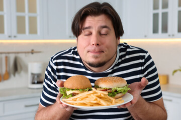 Happy overweight man holding plate with tasty burgers and French fries in kitchen