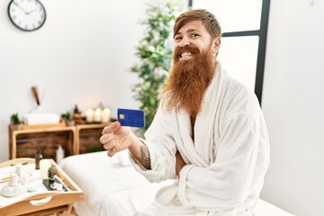 Young redhead man wearing bathrobe holding credit card at beauty center