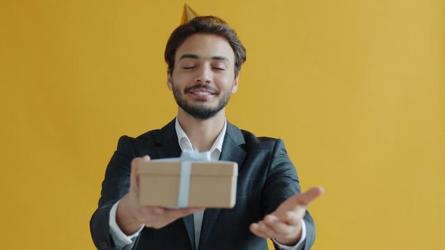 Portrait of generous Arab man giving beautiful gift box stretching hands to camera on yellow background. Birthday and celebration concept.