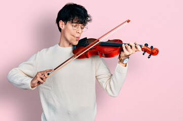 Handsome hipster young man playing violin making fish face with mouth and squinting eyes, crazy and comical.