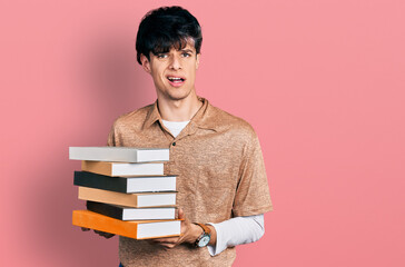 Handsome hipster young man holding a pile of books clueless and confused expression. doubt concept.