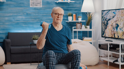 Handheld shot of old man lifting weights and training while sitting on fitness toning ball at home....