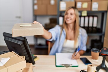 Young blonde woman ecommerce business worker holding order package writing on clipboard at office