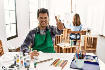 Young artist man at art studio approving doing positive gesture with hand, thumbs up smiling and happy for success. winner gesture.