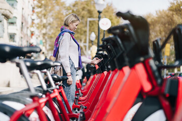 Blond woman taking a red bicycle in a bike rental station in the city with smartphone sharing app -...