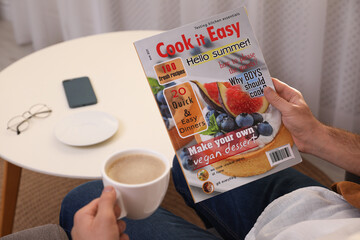 Man with cup of coffee reading magazine in armchair at home, closeup