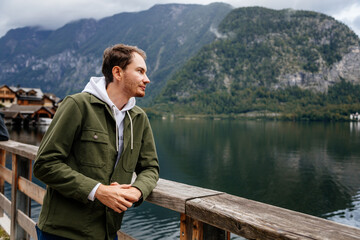 Fototapeta premium Hallstatt, Austria: Portrait of beautiful romantic young man tourist smile, natural light, boy in green jacket, scenic picturesque town street, mountain village near lake, Alps at summer day