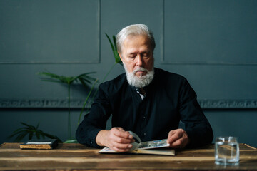 Portrait of serious gray-haired senior adult man looking postage stamps at collection book sitting...