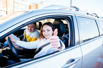 Smiling carefully young woman in drivers seat showing thumbs up gesture of car taking driving...