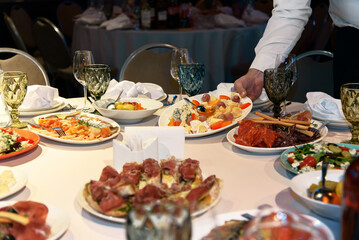 Table is set with variety of gourmet dishes and appetizers for banquet. Waiter's hand putting plate on table. Selective focus.
