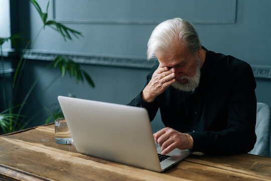 Worried mature gray-haired grandfather grieving, received bad message notification, sitting alone at home. Depressed bearded senior aged man crying looking at laptop computer screen in dark room.