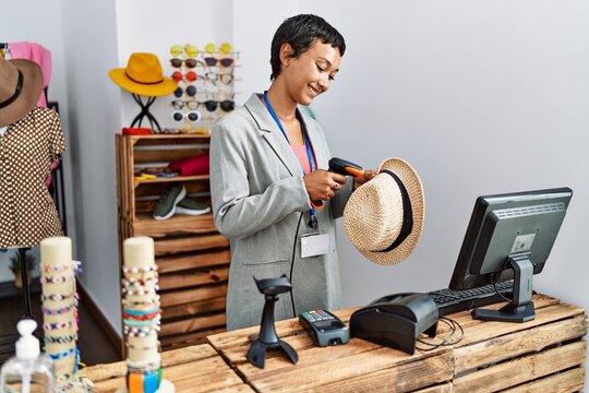 Young hispanic woman shopkeeper smiling confident scanning hat using bardcode reader at clothing store