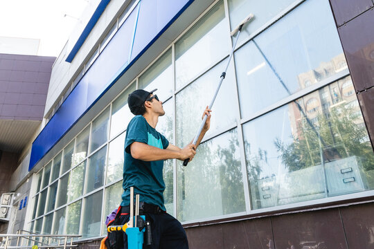 Male Professional Cleaning Service Worker In Overalls Cleans The Windows And Shop Windows Of A Store With Special Equipment