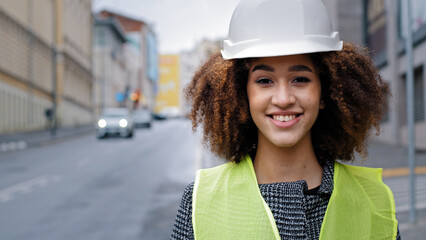 Female portrait worker profession close-up african american woman girl with curly hair civil engineer professional wearing safety helmet standing in city street and looking at camera smiles friendly