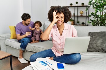 Mother of interracial family working using computer laptop at home doing ok gesture with hand smiling, eye looking through fingers with happy face.