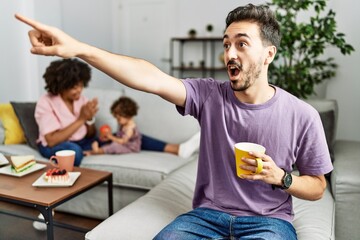 Hispanic father of interracial family drinking a cup coffee pointing with finger surprised ahead, open mouth amazed expression, something on the front
