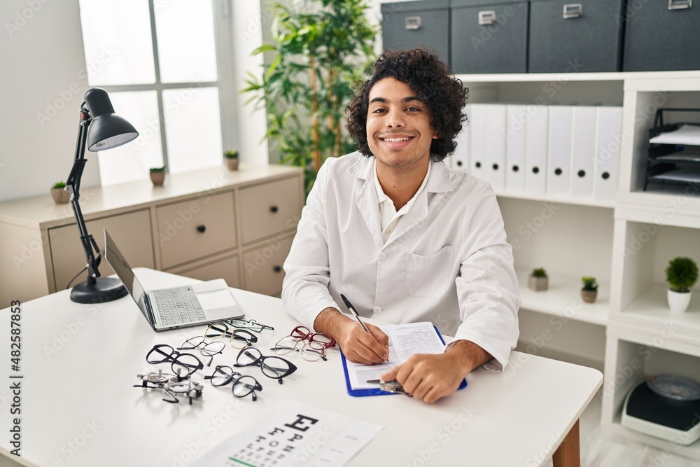 Poster young hispanic man optician writing on clipboard at clinic