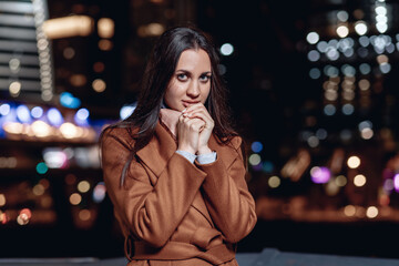 Close up portrait of a lovely pleasant dark-haired lady in a coat posing against the backdrop of the bright lights of the night city looking at the camera from the belts. Female beauty concept