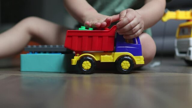 caucasian preschooler boy kid playing with toy car truck and colorful plastic bricks on wooden floor in the living room at home. 3 year child in green shirt creative game box and cubes. educational 