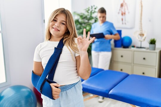 Blonde little girl wearing arm on sling at rehabilitation clinic doing ok sign with fingers, smiling friendly gesturing excellent symbol