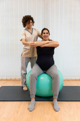 Obraz premium A pregnant woman doing exercises sitting on a fitness ball with her arms in front with the help of physiotherapist