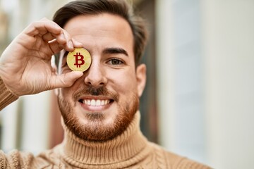 Young caucasian man smiling happy holding bitcoin over eye at the city. - Powered by Adobe