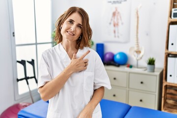 Fototapeta premium Middle age physiotherapist woman working at pain recovery clinic cheerful with a smile of face pointing with hand and finger up to the side with happy and natural expression on face