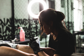 Obraz premium A professional tattoo artist makes a black and white tattoo on a woman's leg, applying in ink. The process of tattooing the skin.