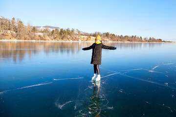 A young athletic girl is skating on the transparent ice of the frozen Lake Baikal on a sunny winter...