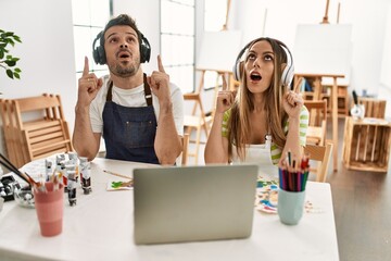 Young couple of wife and husband at art studio looking at video on laptop amazed and surprised looking up and pointing with fingers and raised arms.