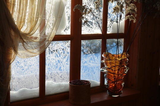 Still life of a winter wooden window in the snow with a pattern on frozen glass with red berries and dried flowers in a vase interior of the home
