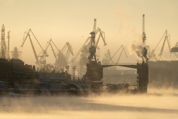 Fototapeta premium Cranes of Baltic shipyard in St. Petersburg in frosty winter day, steam over the Neva river, smooth surface of the river at sunset. 