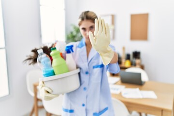 Young blonde woman wearing cleaner uniform holding cleaning products doing stop sing with palm of the hand. warning expression with negative and serious gesture on the face.