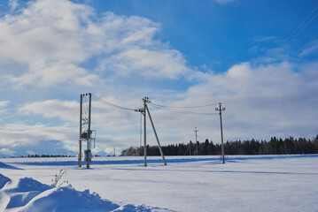 Electric poles on a field covered with snow. Black wires against a bright blue cloudy sky.