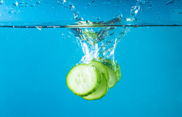 Falling of fresh cucumber slices into water against blue background