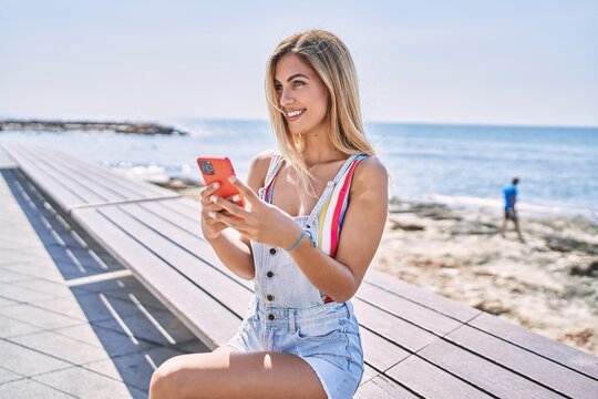 Young blonde girl smiling happy using smartphone sitting on the bench at the beach.