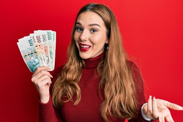 Young blonde woman holding czech koruna banknotes celebrating achievement with happy smile and winner expression with raised hand