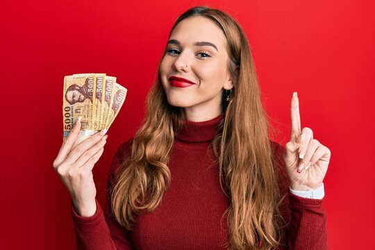 Young blonde woman holding 5000 hungarian forint banknotes smiling with an idea or question pointing finger with happy face, number one