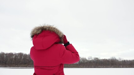 Woman puts on warm hood of winter coat standing outdoor in white snowy frosty countryside landscape background