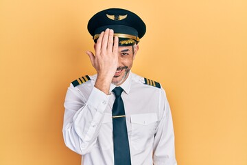 Handsome middle age man with grey hair wearing airplane pilot uniform covering one eye with hand, confident smile on face and surprise emotion.