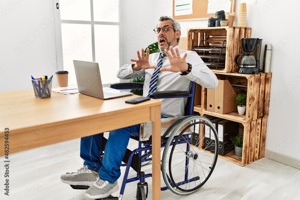 Canvas Prints Middle age hispanic man working at the office sitting on wheelchair afraid and terrified with fear expression stop gesture with hands, shouting in shock. panic concept.