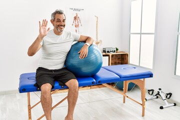 Middle age hispanic man at pain recovery clinic holding pilates ball waiving saying hello happy and...